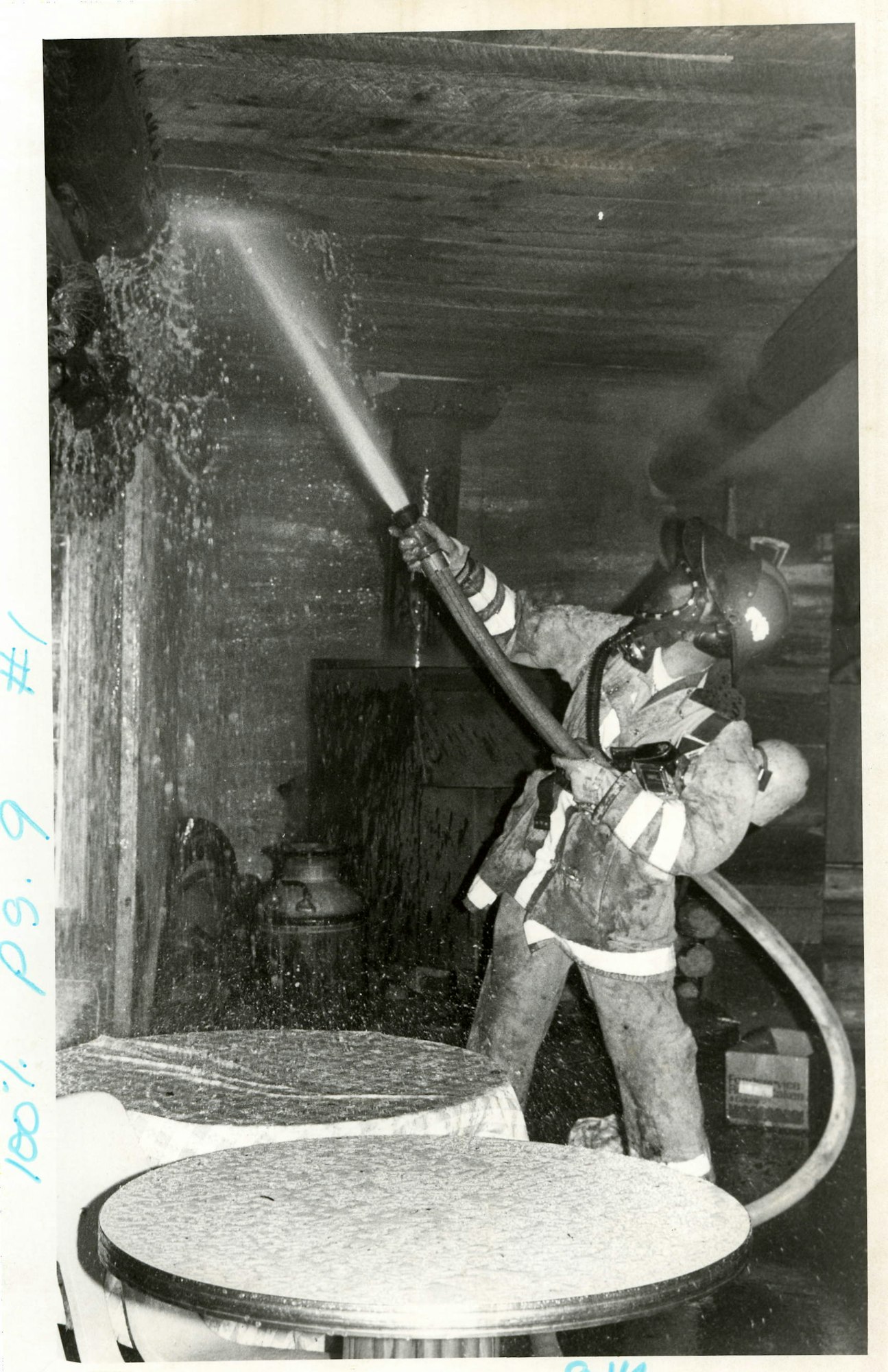 A firefighter using a hose, likely extinguishing a fire; water spray visible, in a building with furniture.