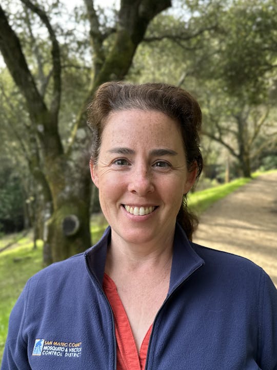 A person smiling outdoors wearing a blue jacket with a "San Mateo County Mosquito & Vector Control District" logo.