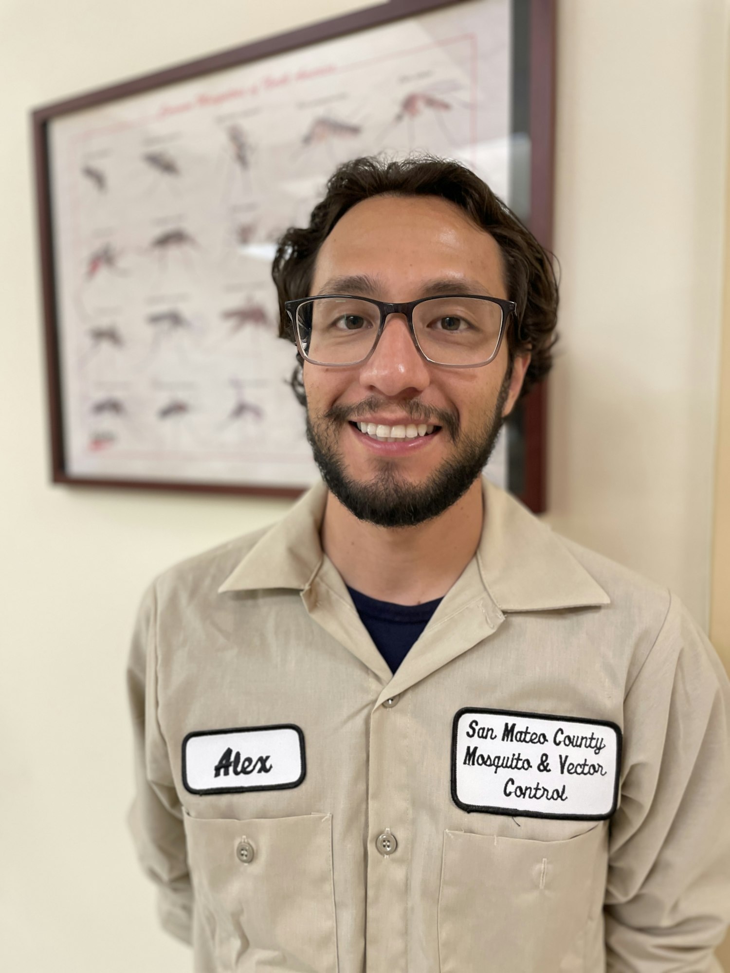 A person in a beige uniform with name tags, standing in front of a mosquito chart.