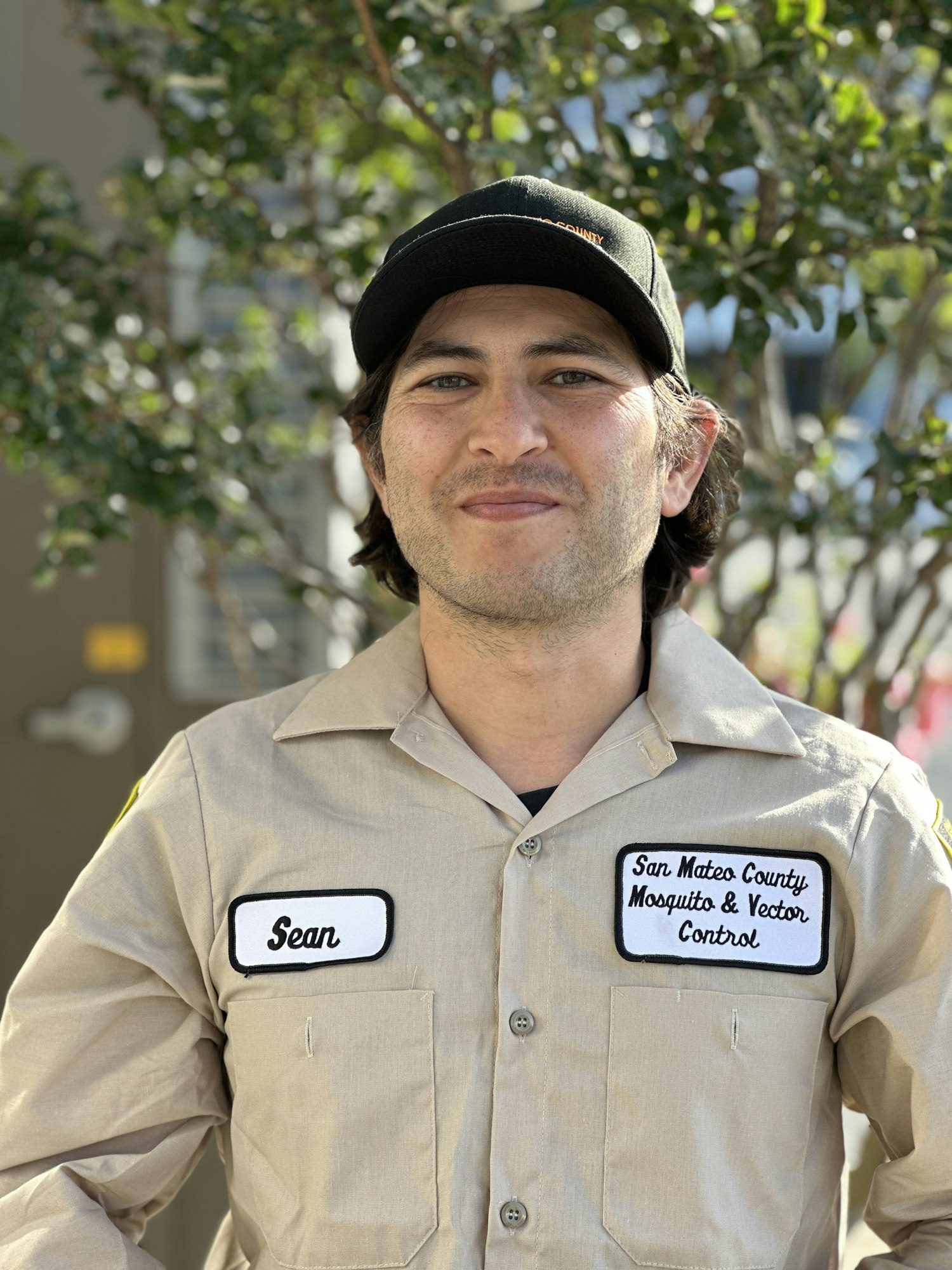 A man in a uniform with name tag "Sean" and patch of "San Mateo County Mosquito & Vector Control" with trees in the background.