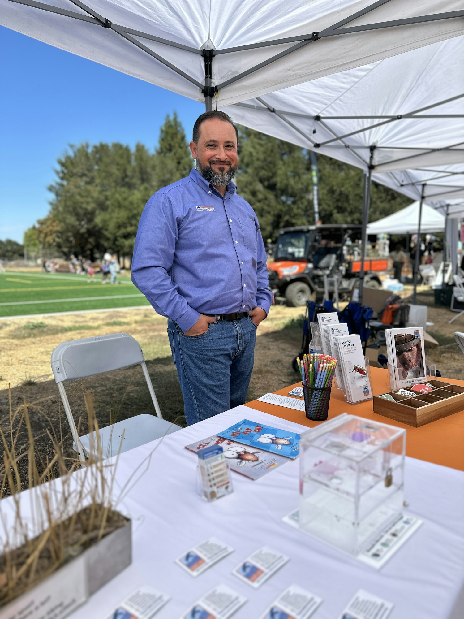 Man in blue shirt standing at a booth under a canopy with brochures, pencils, and display items outdoors.