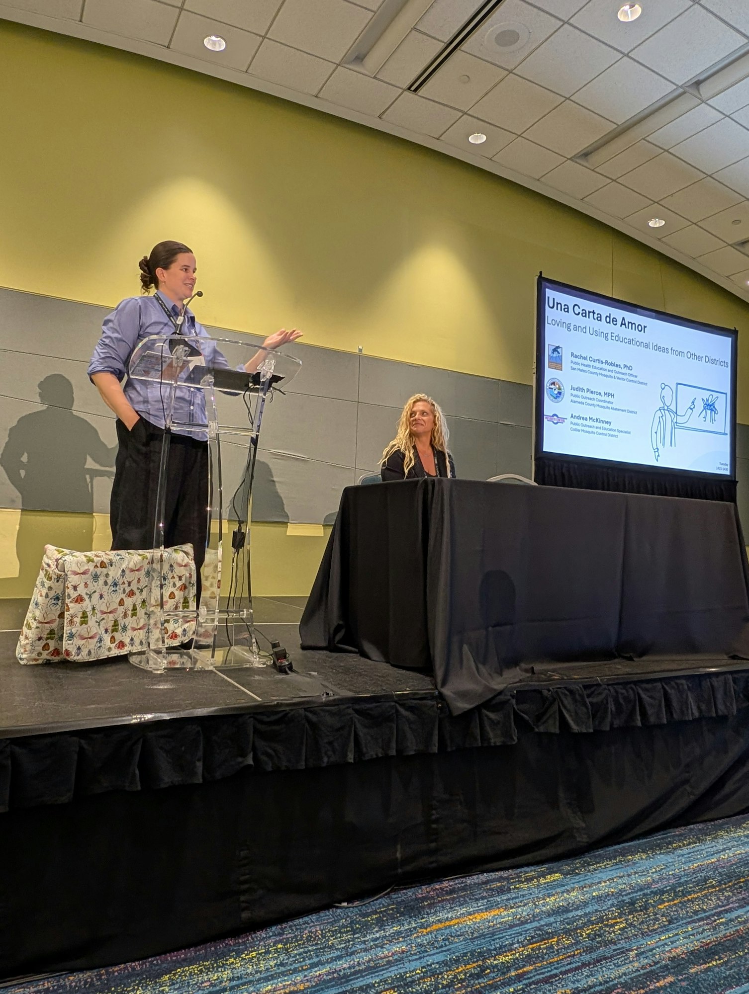 A person speaks at a podium while another sits at a table. A slide titled "Una Carta de Amor" is projected on the screen.
