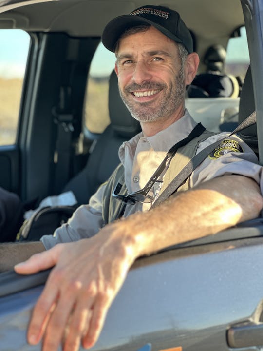 A man smiling while sitting in a vehicle, wearing a hat and uniform that reads "San Mateo County Mosquito & Vector Control District."