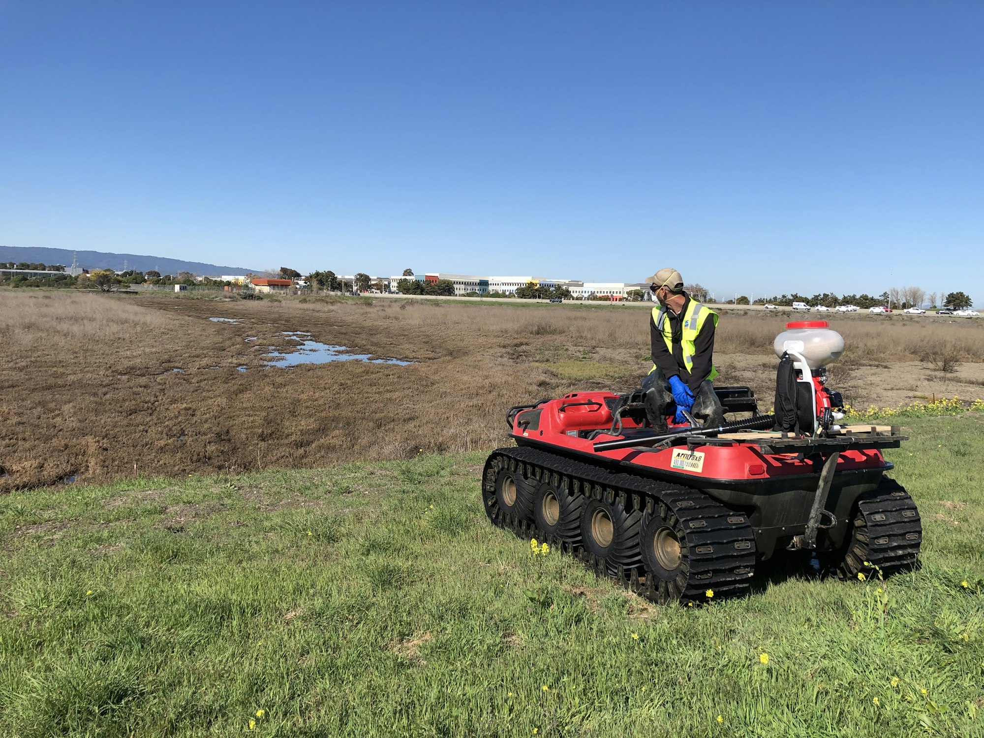 Photograph of a landscape that is green grass in the front and brown marsh in the back. On the green grass, there is a machine vehicle that has treaded wheels and a person sitting on top looking back at the brown marsh.