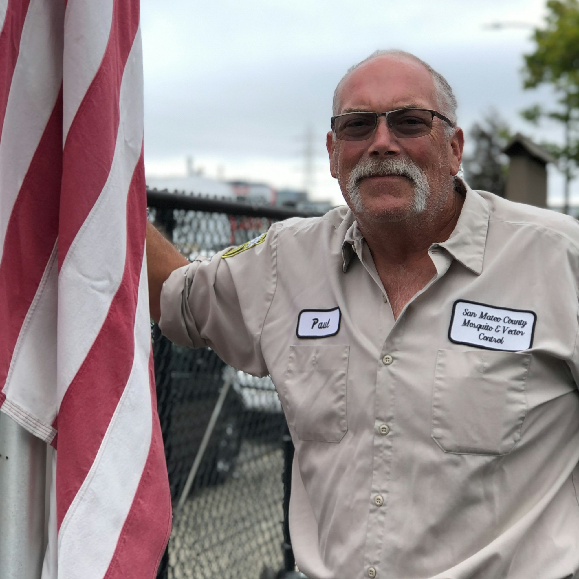 A person with dark glasses and a moustache in a tan uniform shirt with the name "Paul" on the name tag. Standing near the red and white stripes of a draped US flag.
