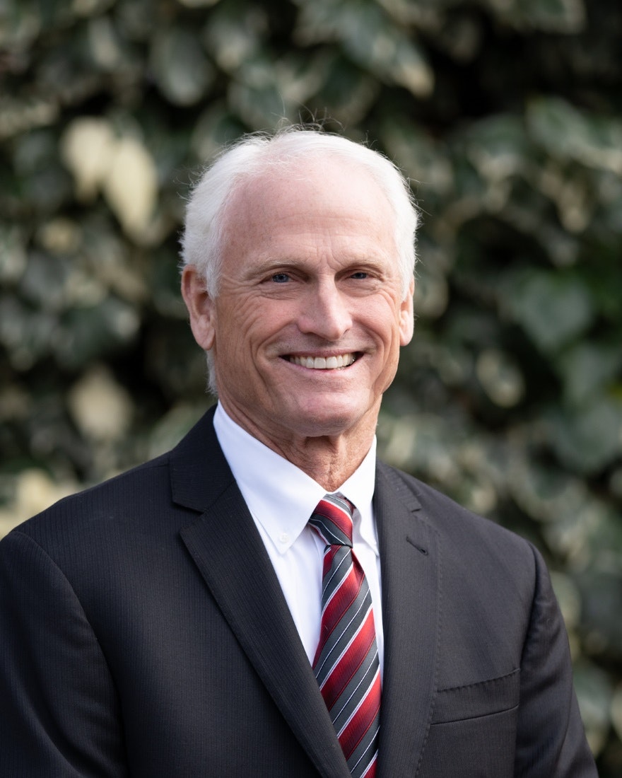 Photograph of person in white collared shirt with striped tie and dark suit jacket. The person has short white hair and is smiling.