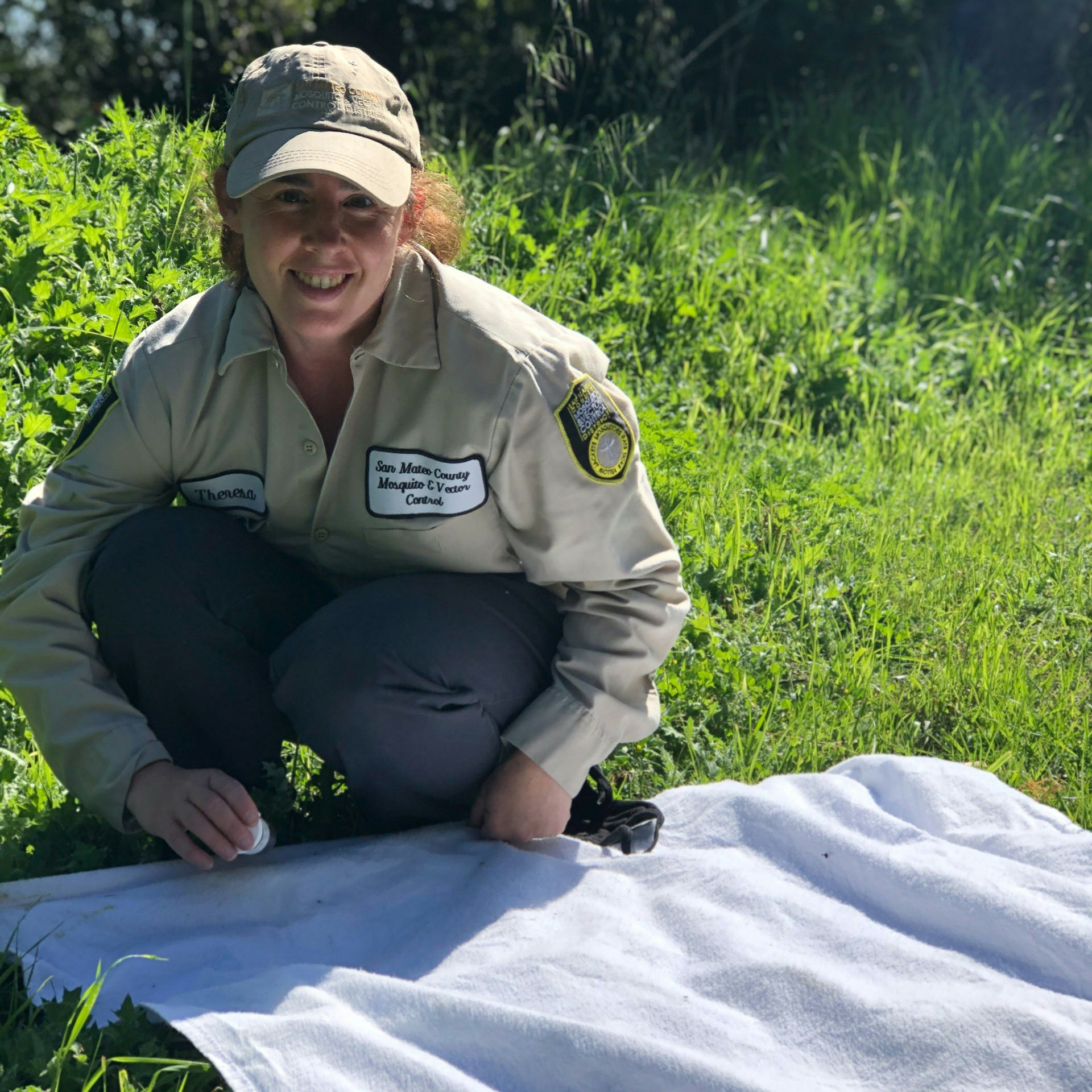 Person in tan uniform and hat squatting on ground with green grass in the background and a white cloth in front.