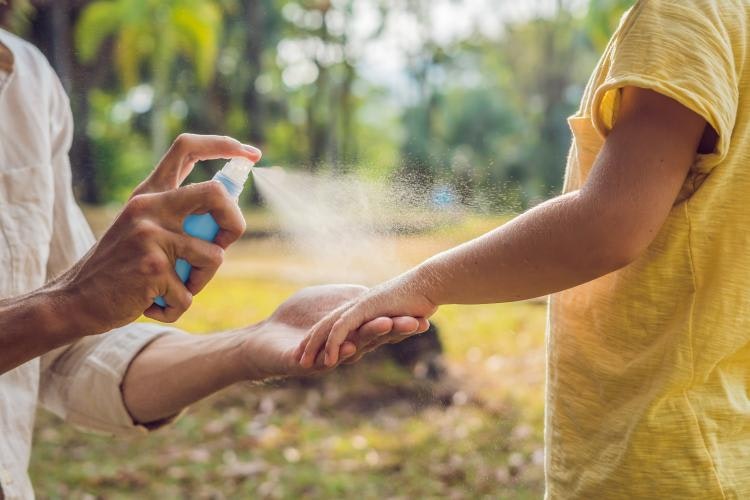 Side view of an adult and a child. The adult is holding a spray bottle and spraying the arm of the child.  The child has a yellow short-sleeve shirt.