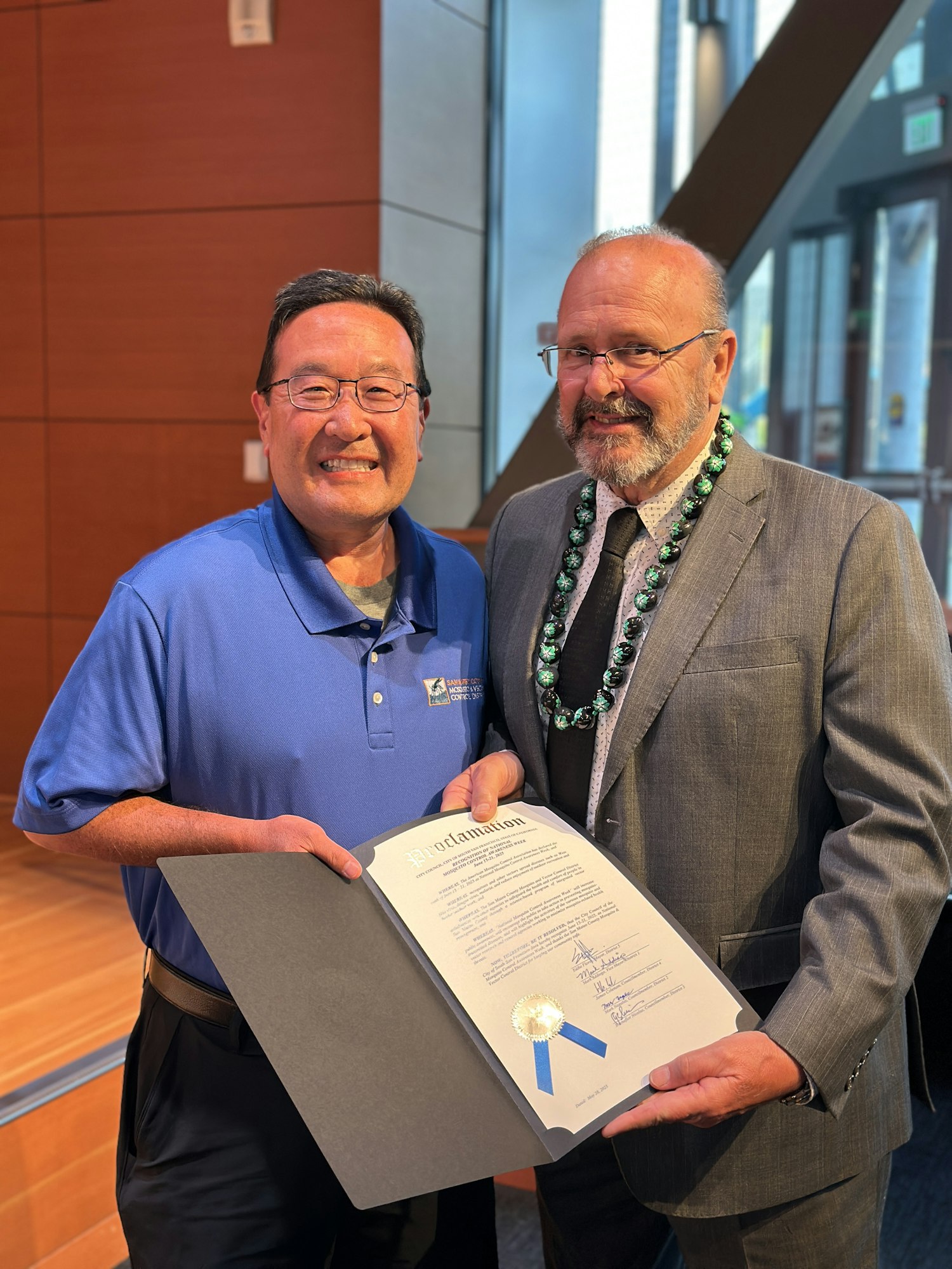 Two men are smiling and holding a framed proclamation document indoors.