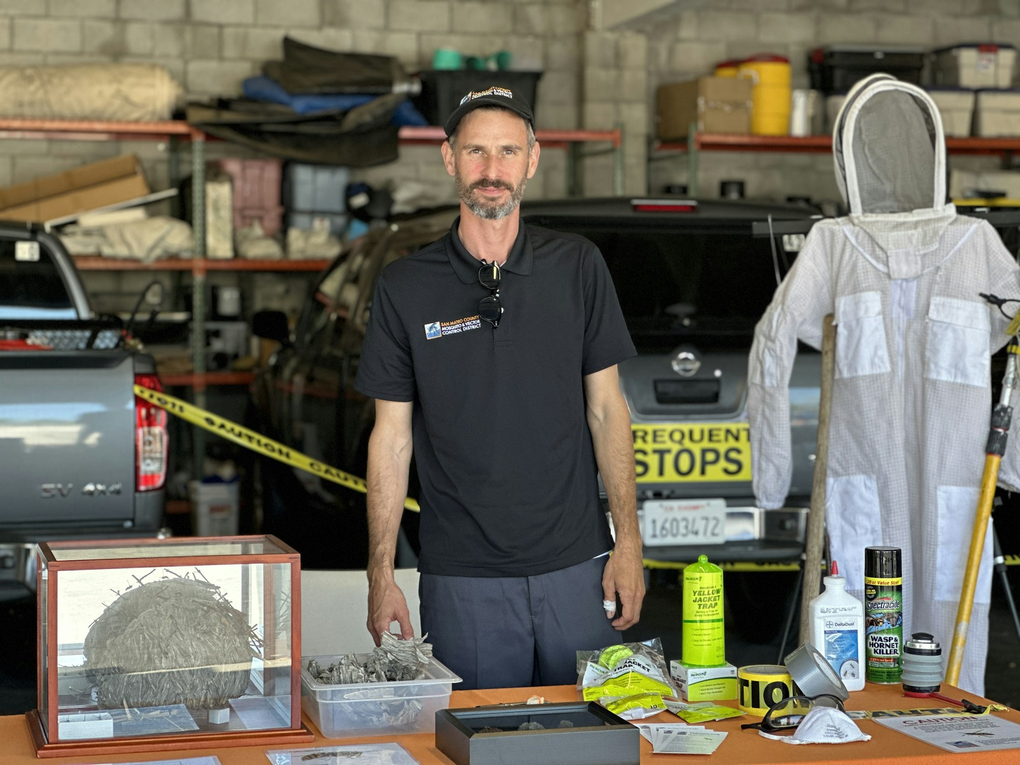 Photograph of a person standing behind a table full of items, including a Yellowjacket next, a bright yellow trap, and a protective 'bee suit'