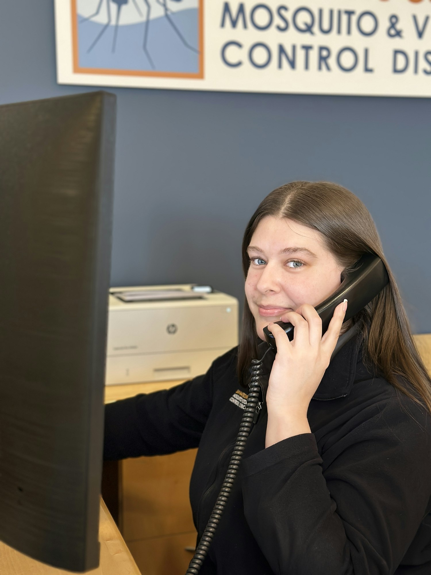 A woman at a desk on the phone with a sign reading "Mosquito & Vector Control District" in the background.