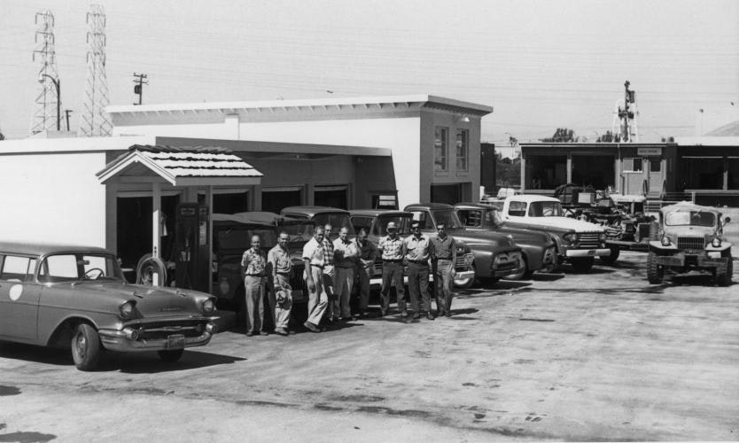 Grey and white photo of several buildings with vehicles from the mid-1900s and several people facing the camera in the parking lot