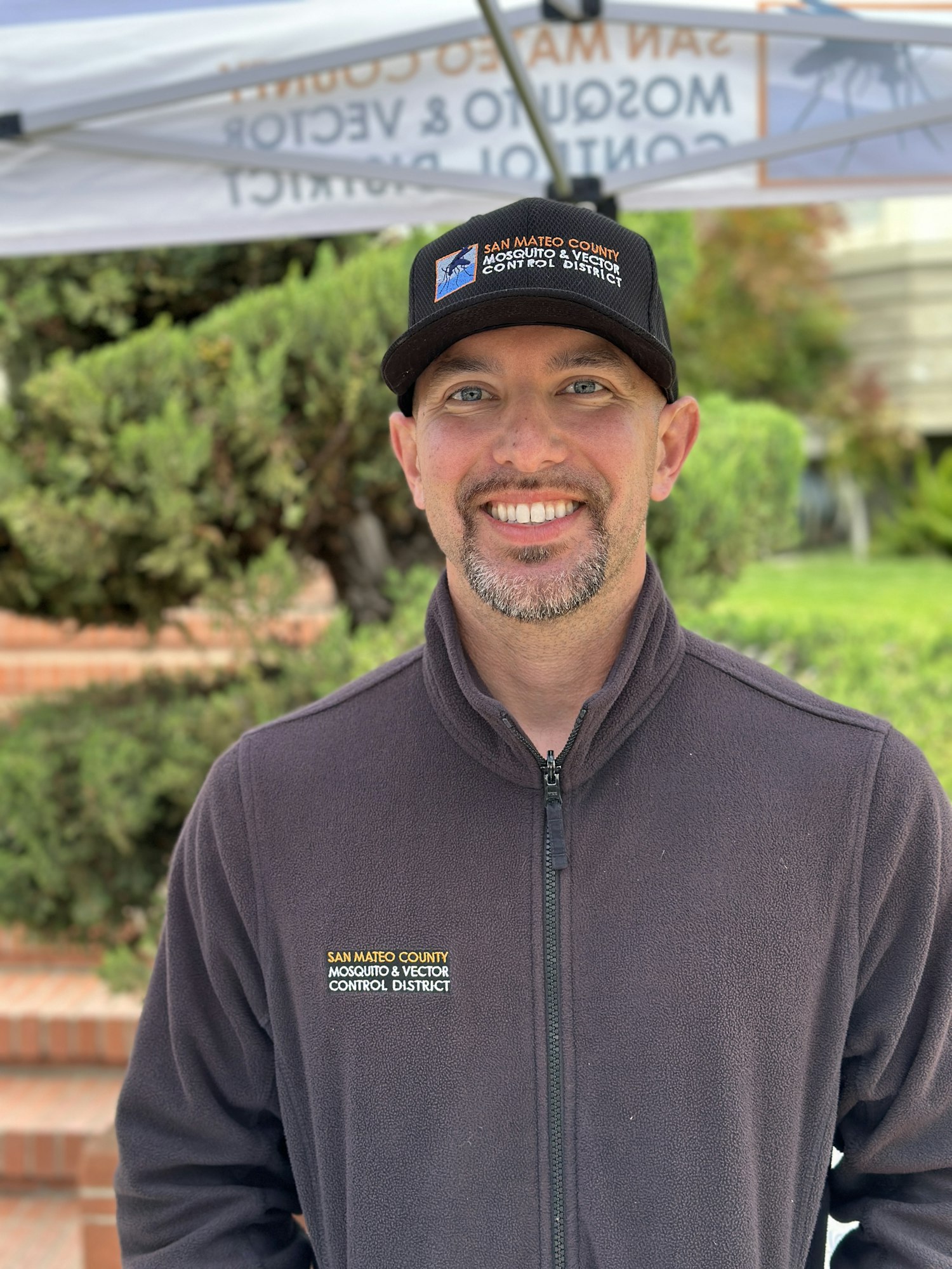 A person smiling, wearing a "San Mateo County Mosquito & Vector Control District" jacket and cap, standing outdoors.
