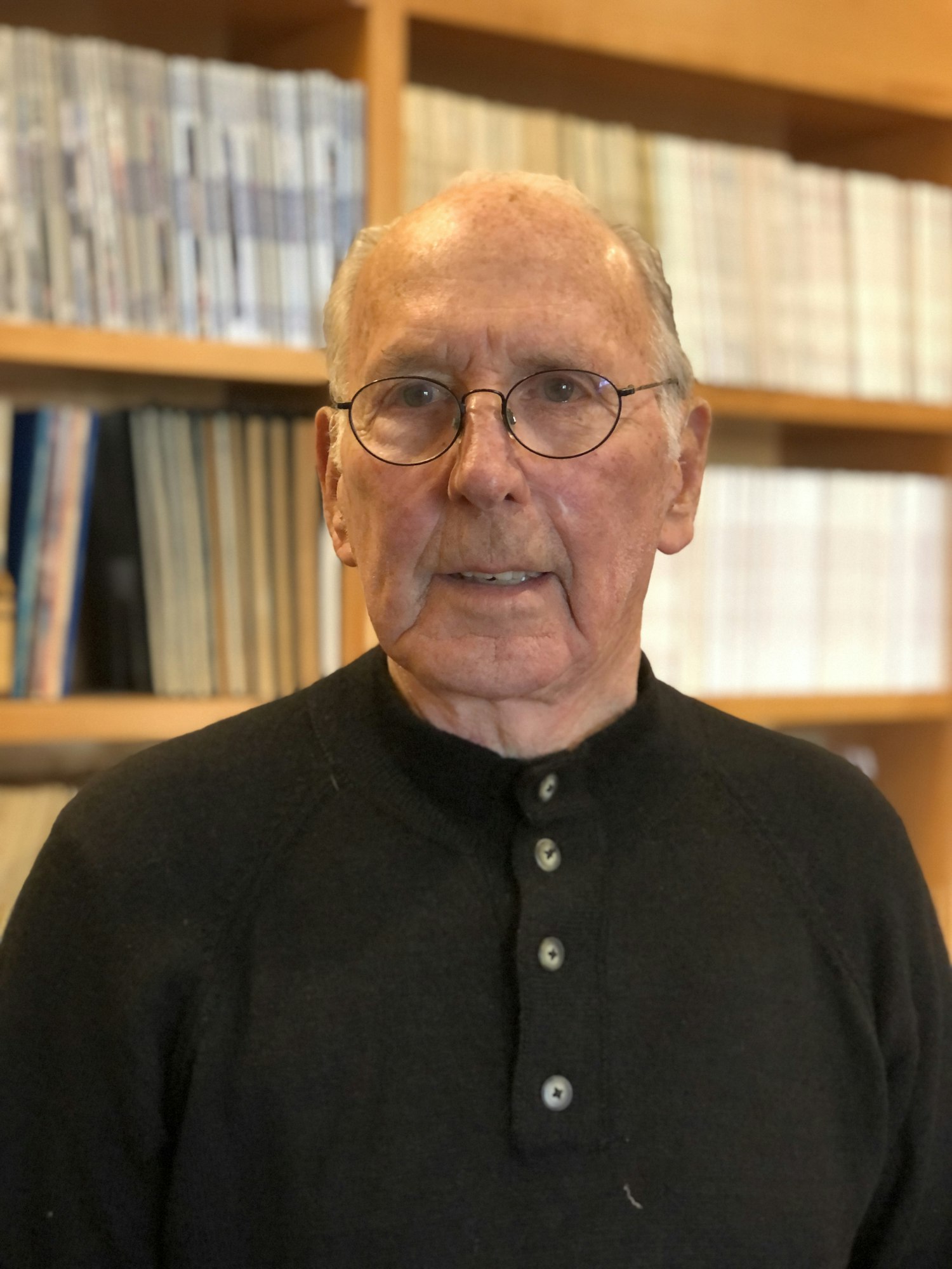 Photo of man in glasses and black shirt looking at camera, background is rows of books