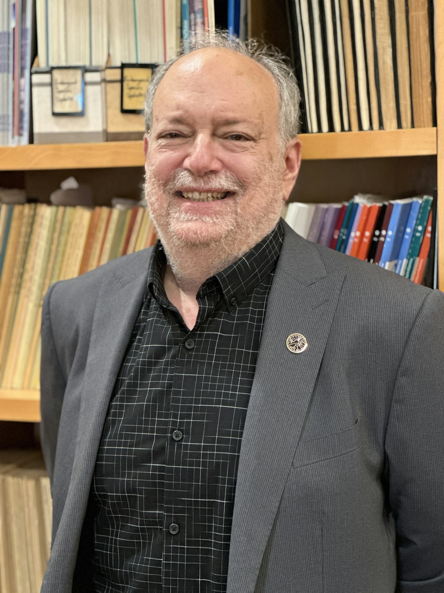 A smiling person in a suit with badge, standing in front of a bookshelf with various books and folders.