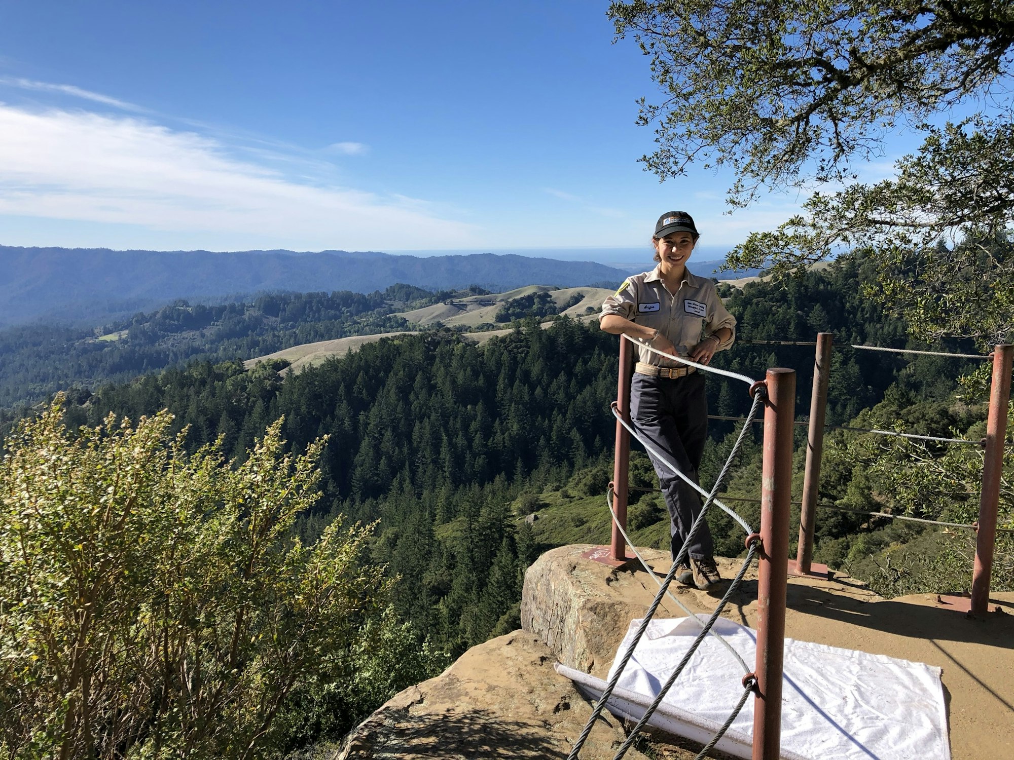 A person in a uniform stands on a rock platform overlooking a scenic valley and forested hills.