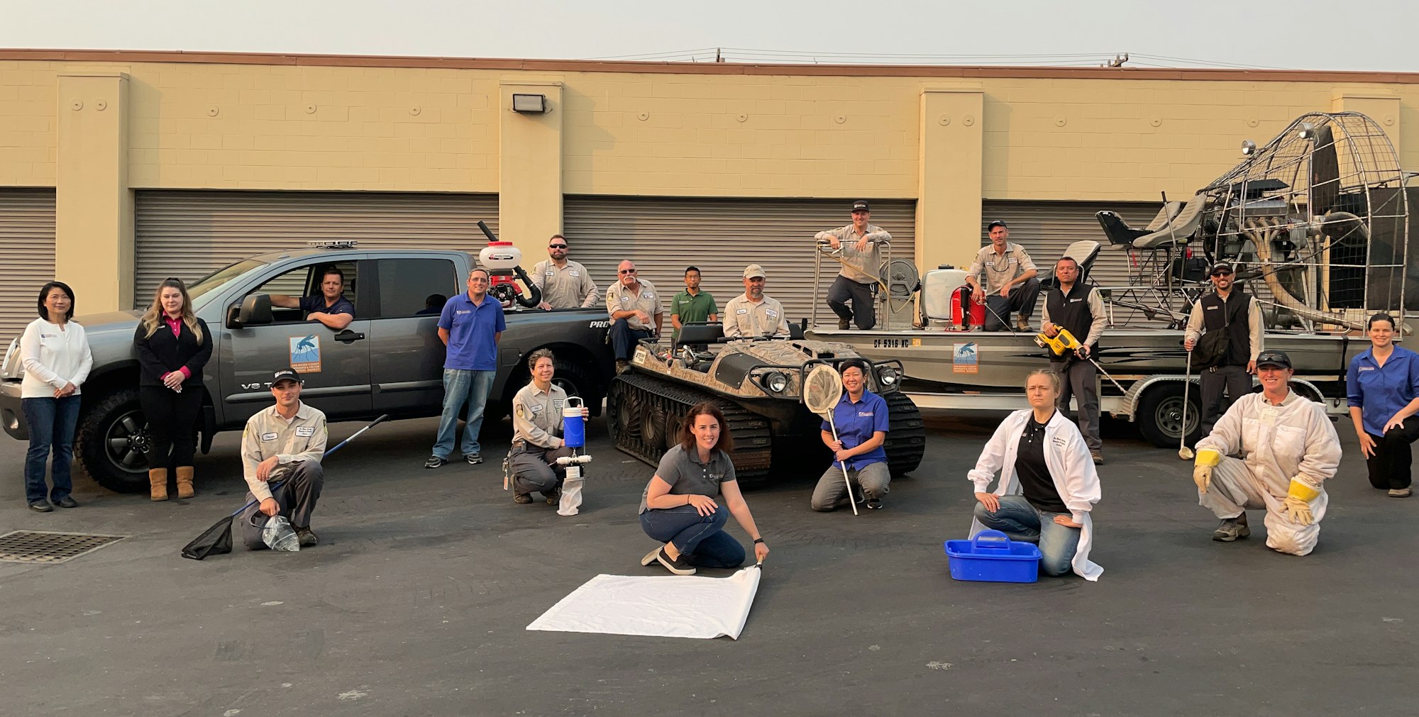 19 people in front of a building. Many are in tan uniforms, some are holding equipment used for vector control. There is a pick-up truck, an Argo (a small vehicle used for accessing marsh areas, and an airboat (has a large fan on the back) on a trailer.