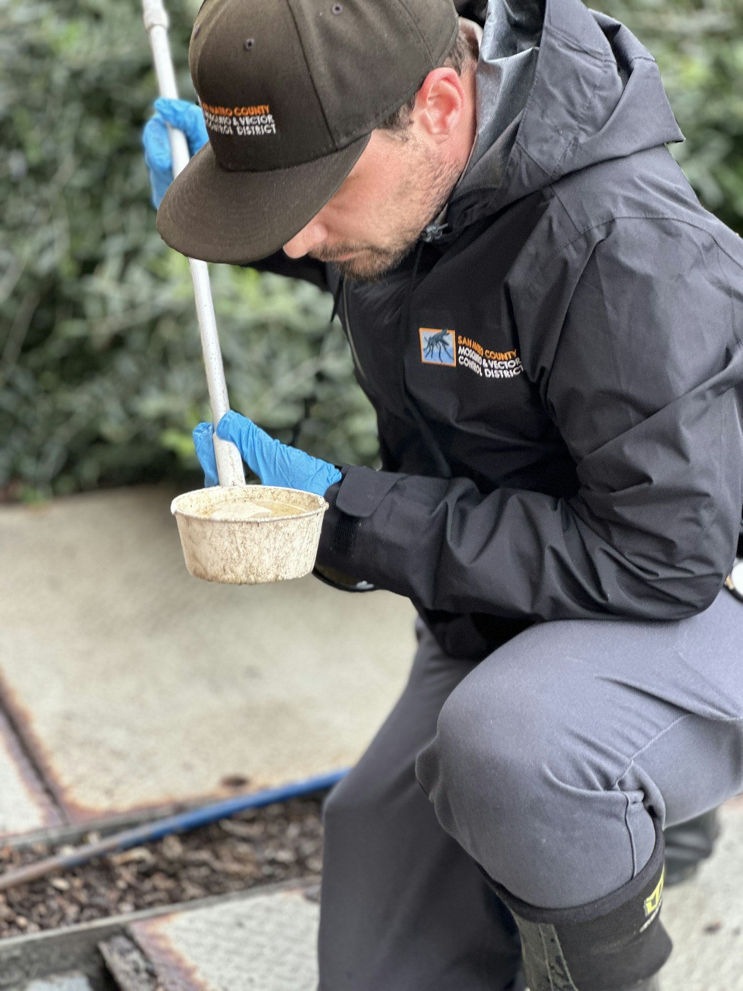 A person in outdoor gear collects a sample using a long-handled tool, wearing blue gloves, with "Mosquito & Vector Control District" on their jacket.