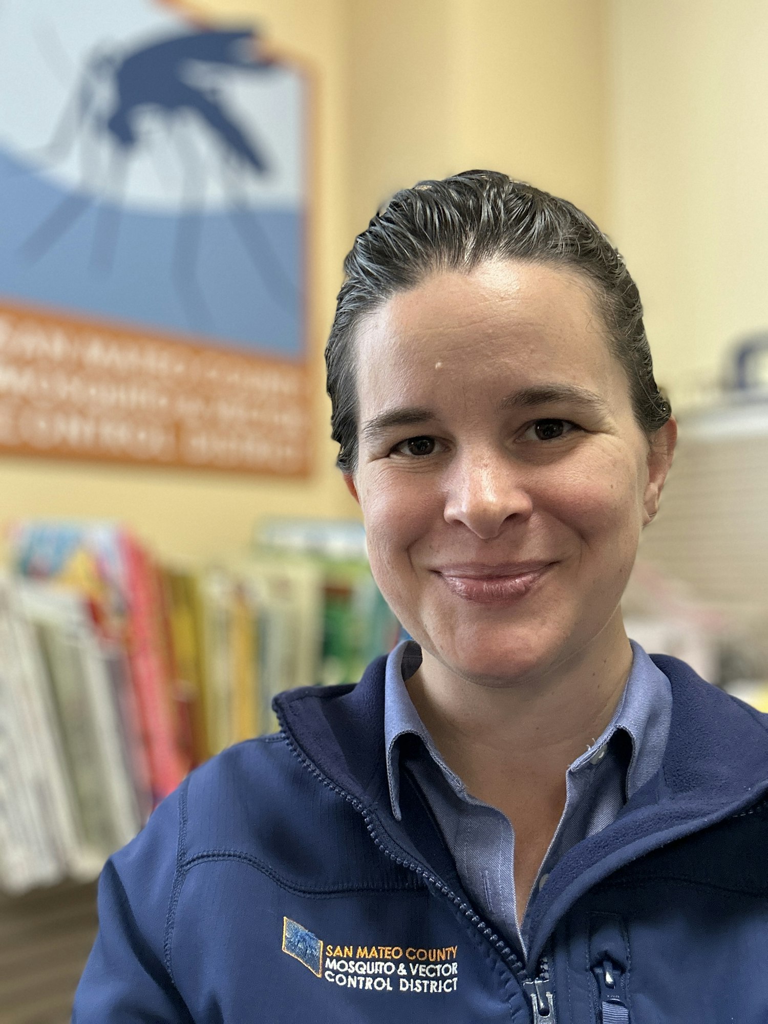 A person in a jacket with "San Mateo County Mosquito & Vector Control District" logo, smiling indoors with a blurred background sign.