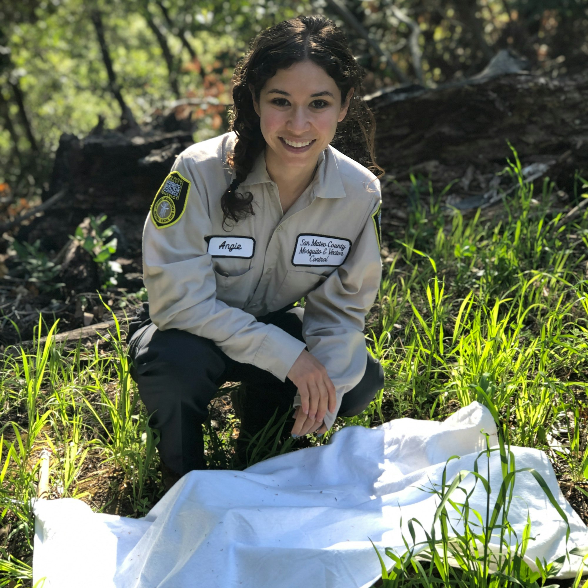 Smiling person with shoulder-length dark hair in tan uniform and hat squatting on ground with green grass in the background and a white cloth in front.
