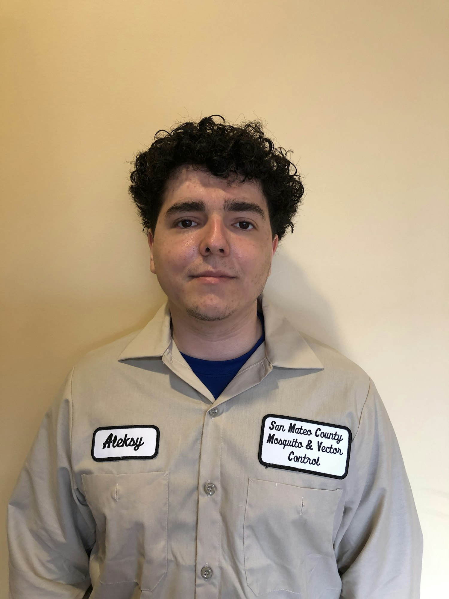 Person in a light brown shirt with patches with the name "Aleksy" and "San Mateo County Mosquito and Vector Control District" Person is standing in front of a tan background.