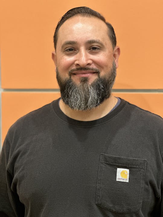 A smiling man with a beard wearing a black T-shirt against an orange background.
