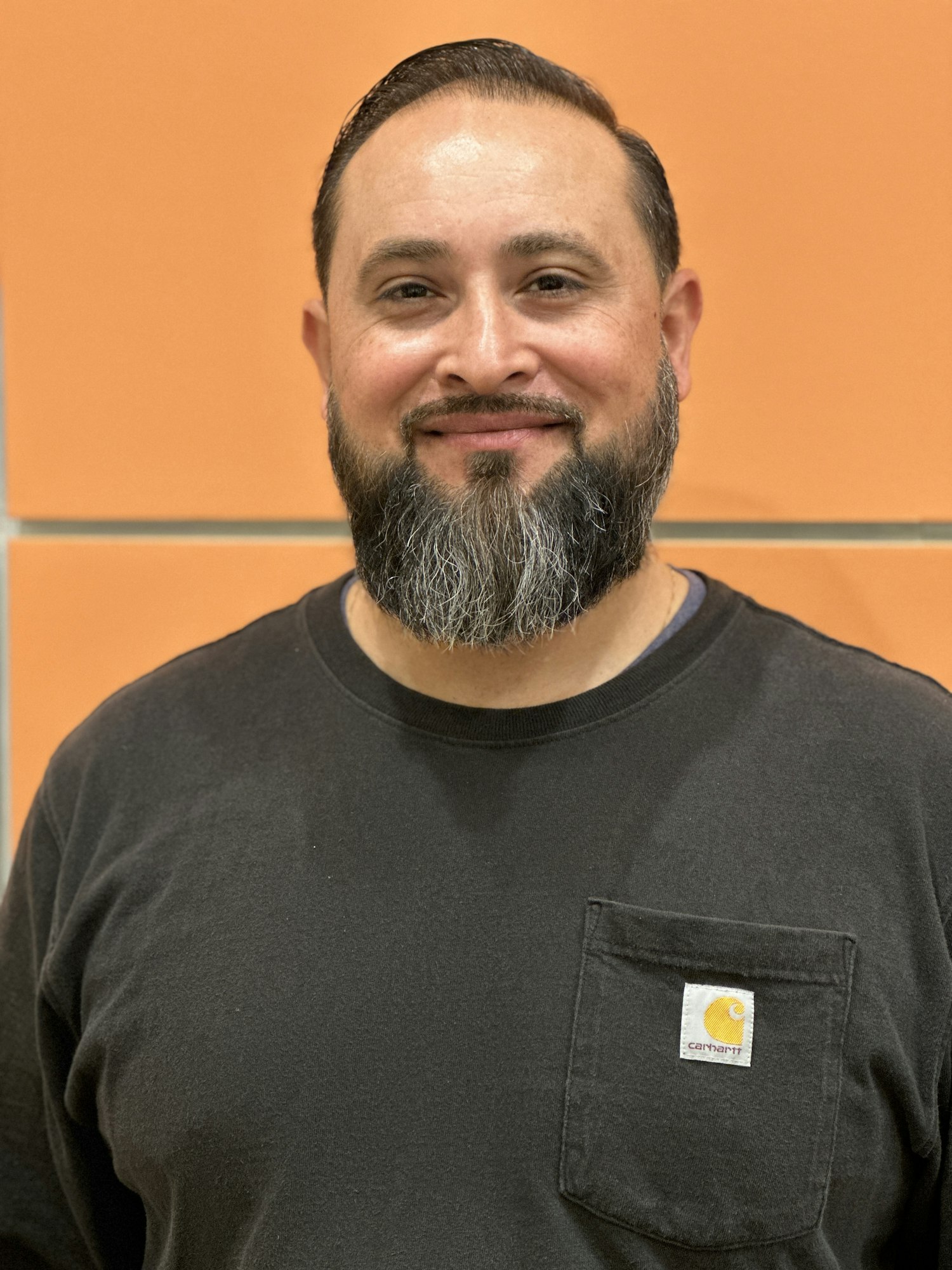 A smiling man with a beard wearing a black T-shirt against an orange background.