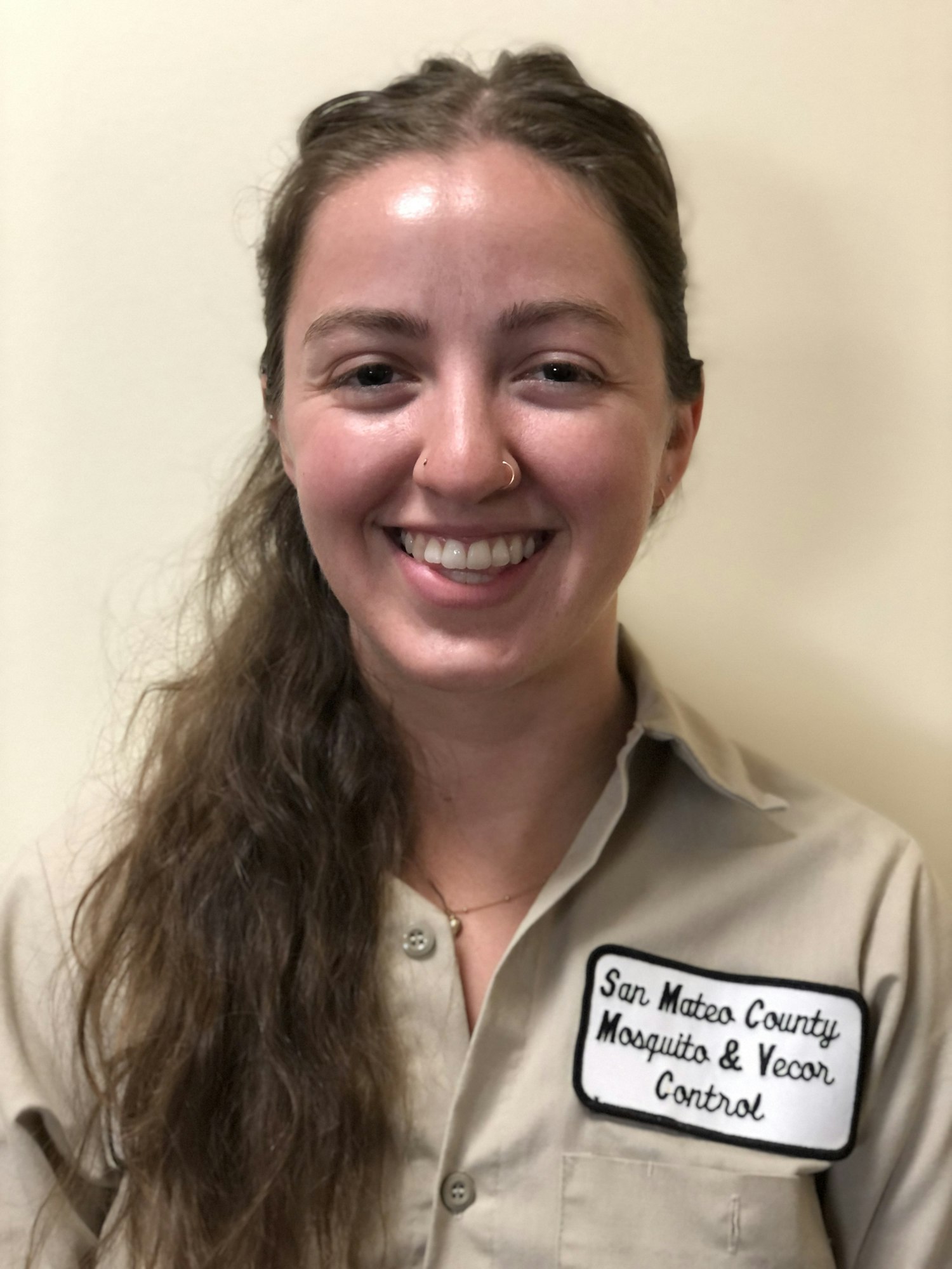 Photo of a smiling person with long brown hair wearing a tan uniform shirt with a label that says 'San Mateo County Mosquito & Vector Control"