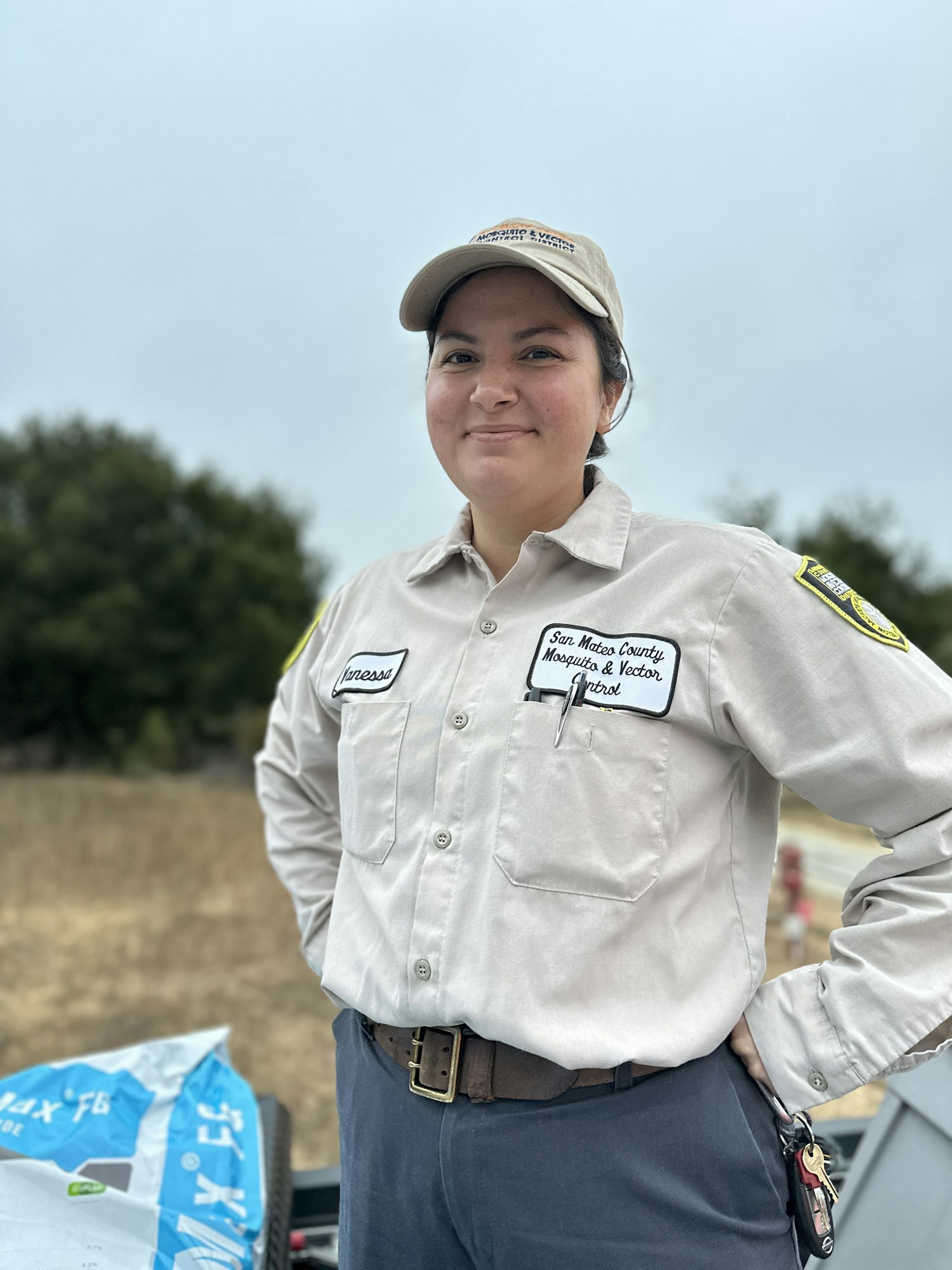 A person in a beige uniform and cap, standing outdoors, with patches and labels on the shirt. Trees and blue sky in the background.