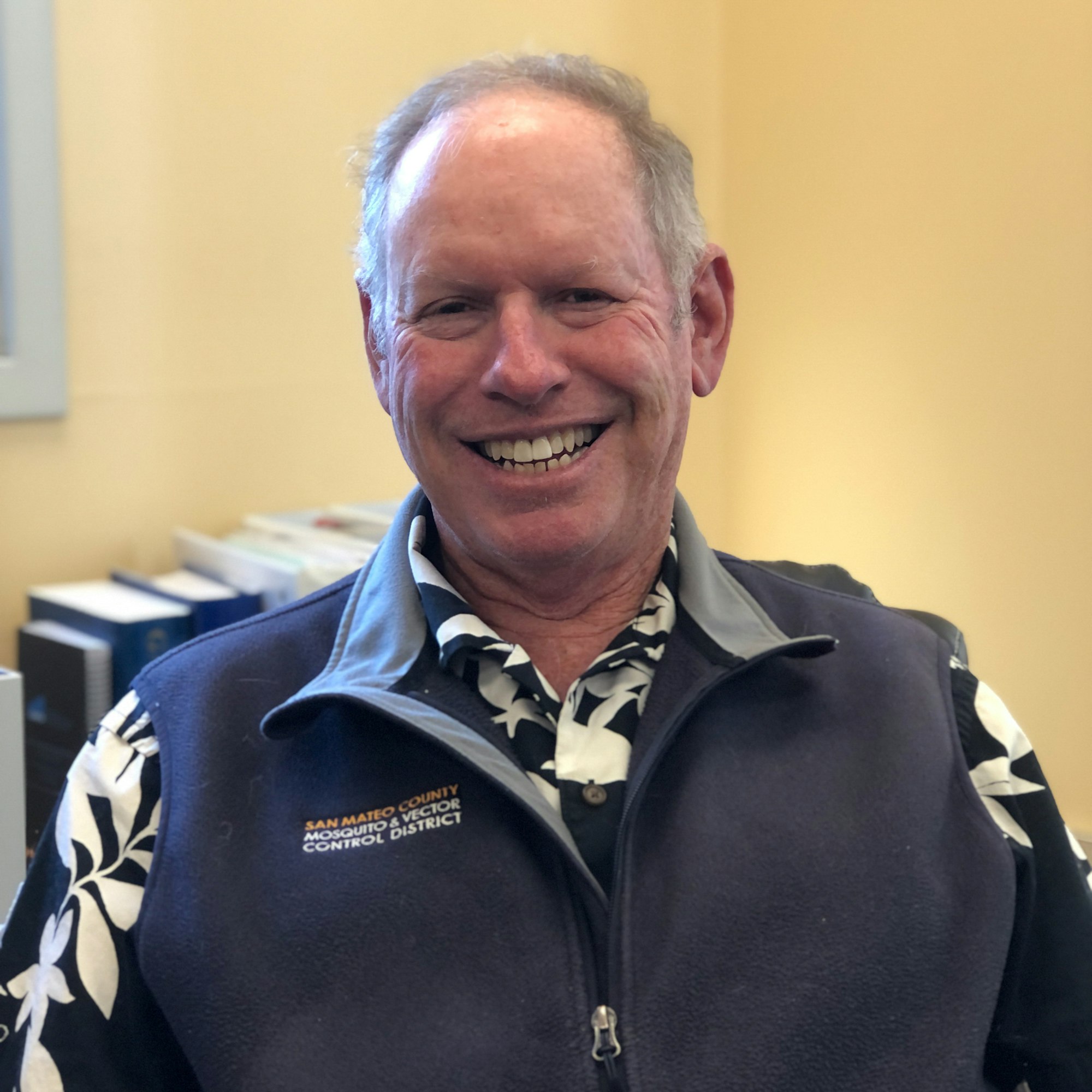 Person smiling with short grey hair in blue and white shirt and dark blue vest with the District logo on it.