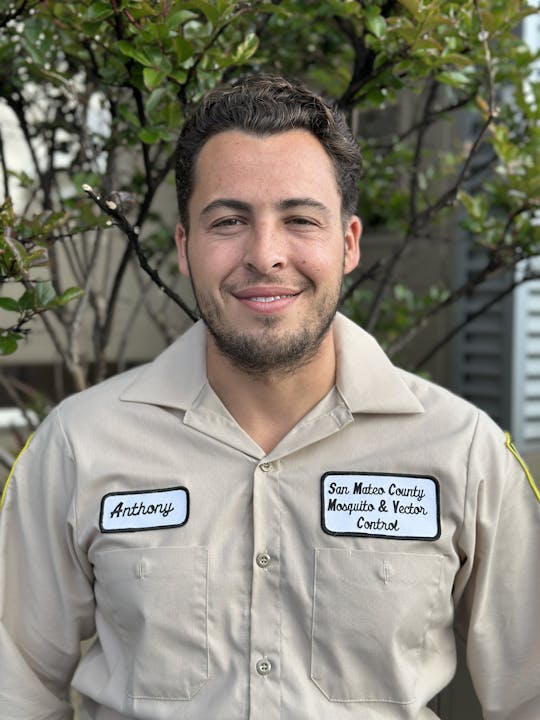 A person wearing a uniform labeled "Anthony" and "San Mateo County Mosquito & Vector Control," standing in front of green bushes.