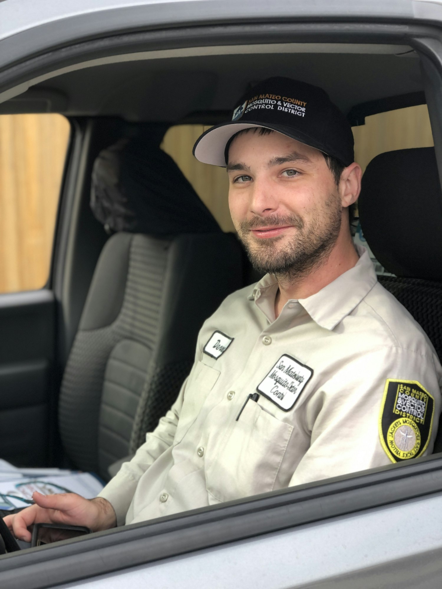 A person in a work uniform sits in a vehicle, wearing a cap with a logo and holding a phone.