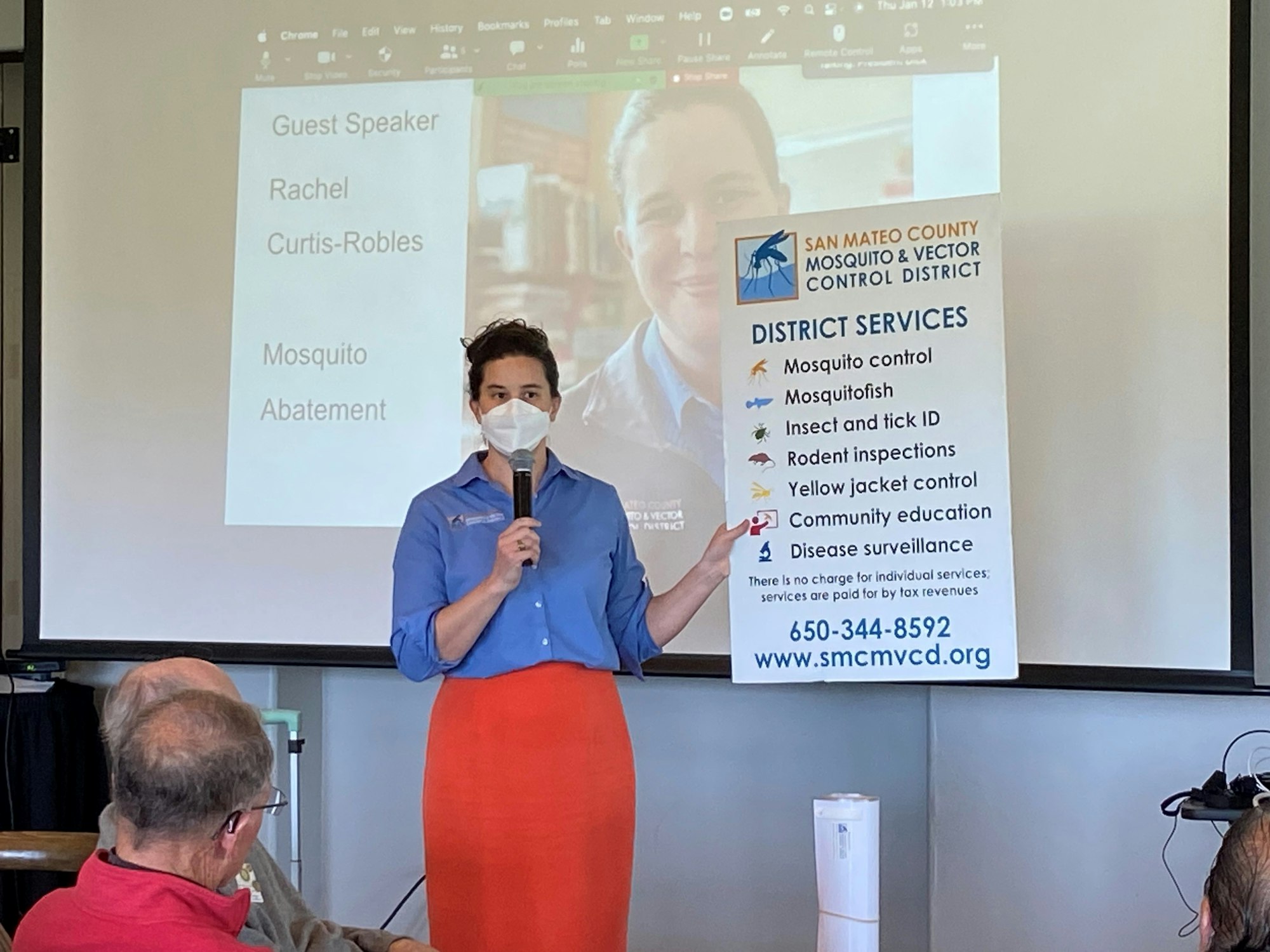 Photo of a person in white mask, blue shirt, and orange skirt holding a microphone and a large poster that says "District Services: Mosquito control, mosquitofish, insect and tick ID, rodent inspections, yellow jacket control, community education, disease surveillance" in front of an audience and powerpoint screen