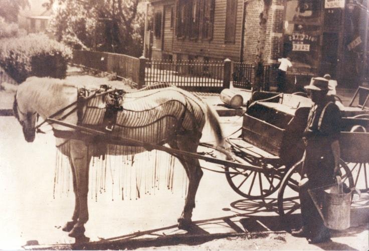 Sepia photograph of a light colored horse hitched to a cart, with a person standing nearby holding a metal pail