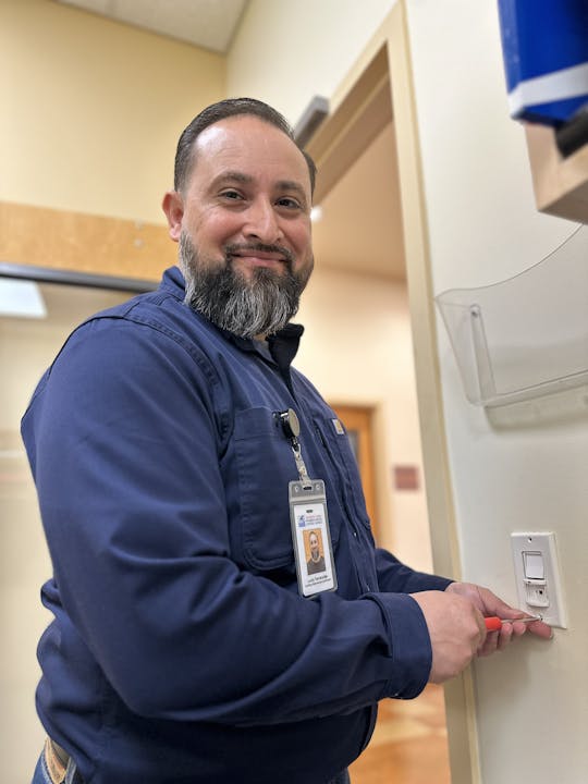 A man with a beard is smiling while using a tool on a light switch, wearing a work uniform and ID badge.