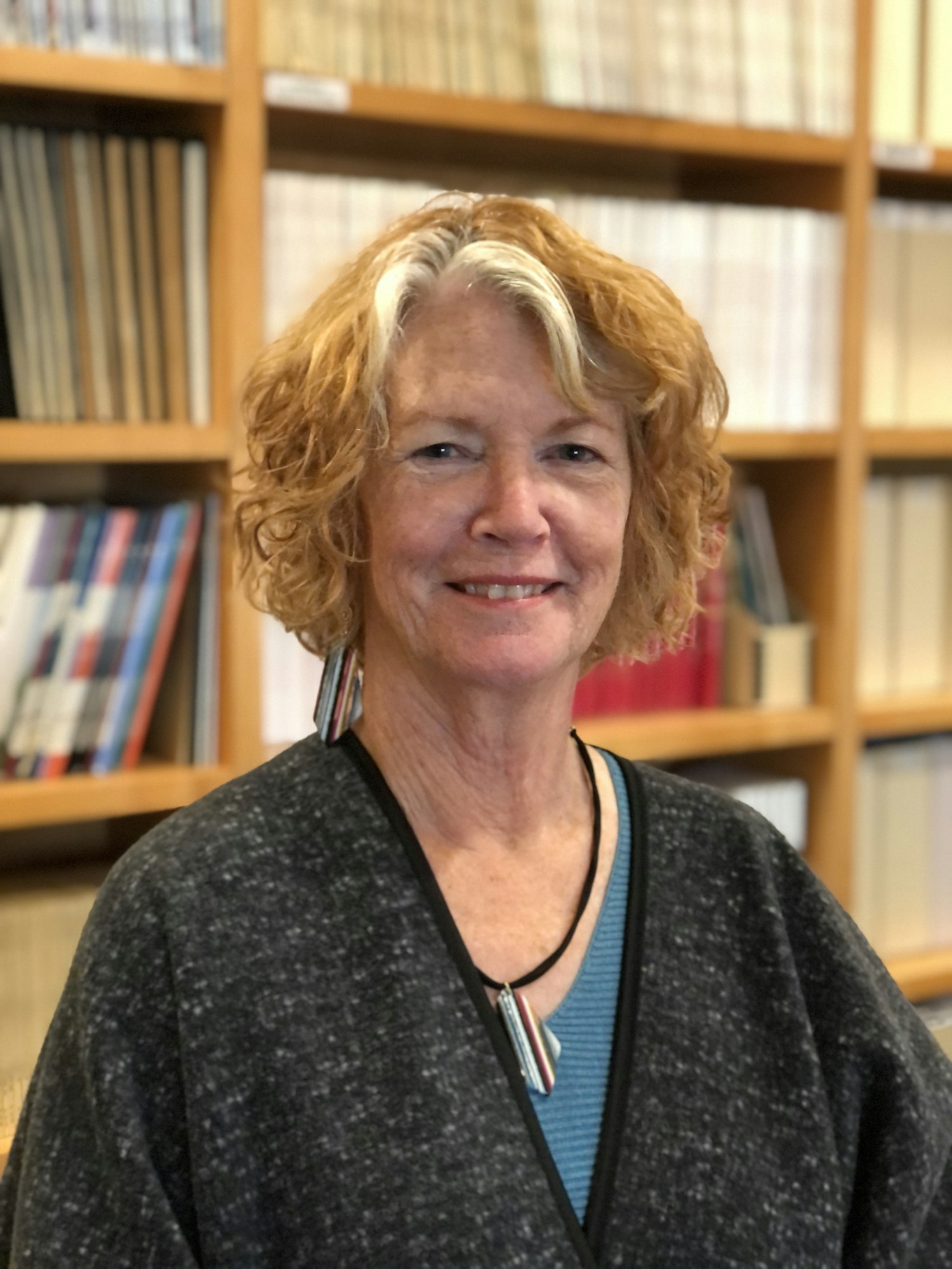 Photograph of smiling woman with short blonde hair, a necklace, and a dark gray top standing in front of rows of books