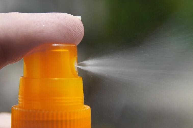 Close-up of a finger tip pressing down on the very top of an orange spray bottle, with a spray coming out of the nozzle.