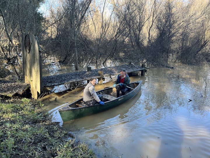 Two people are in a canoe on a flooded, muddy river near a wooden dock, surrounded by leafless trees.