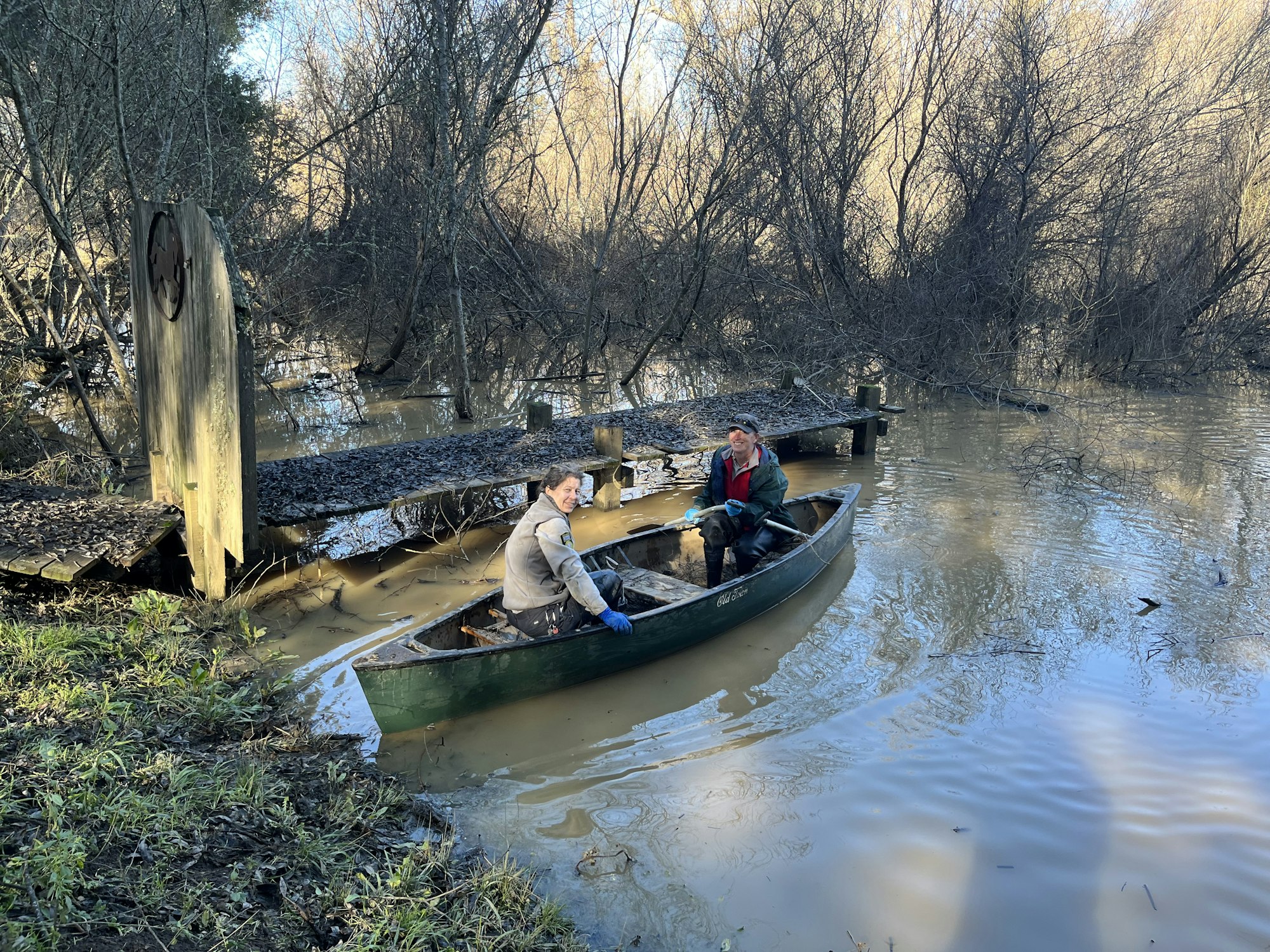 Two people are in a canoe on a flooded, muddy river near a wooden dock, surrounded by leafless trees.