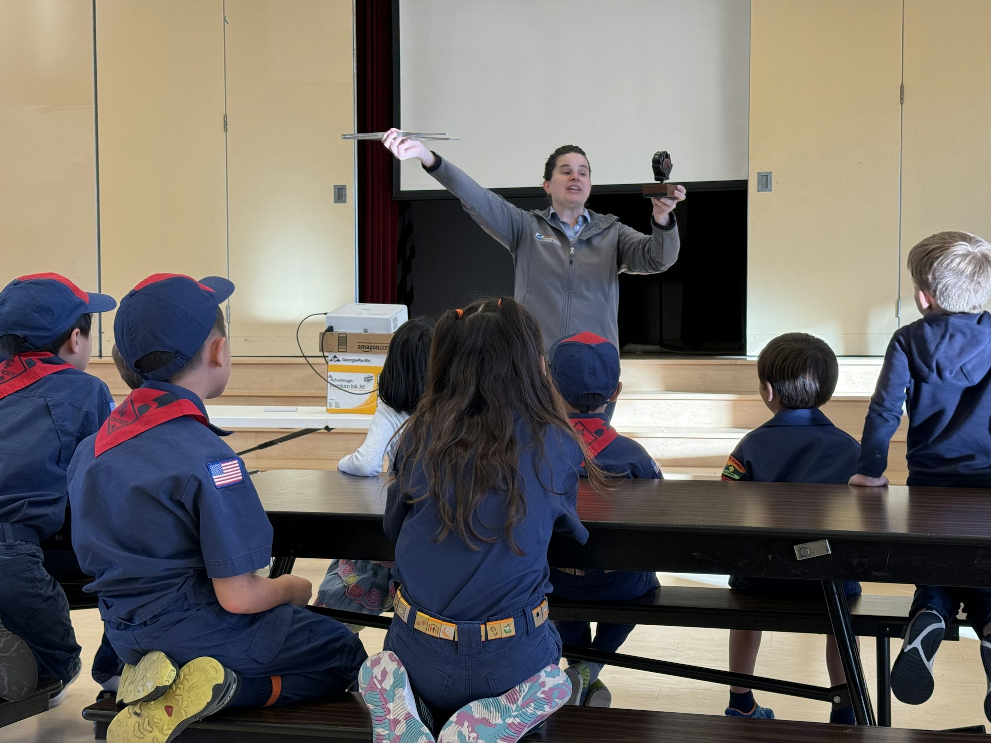 A person presents to a group of kids in uniform, holding an object in a room with a projector.