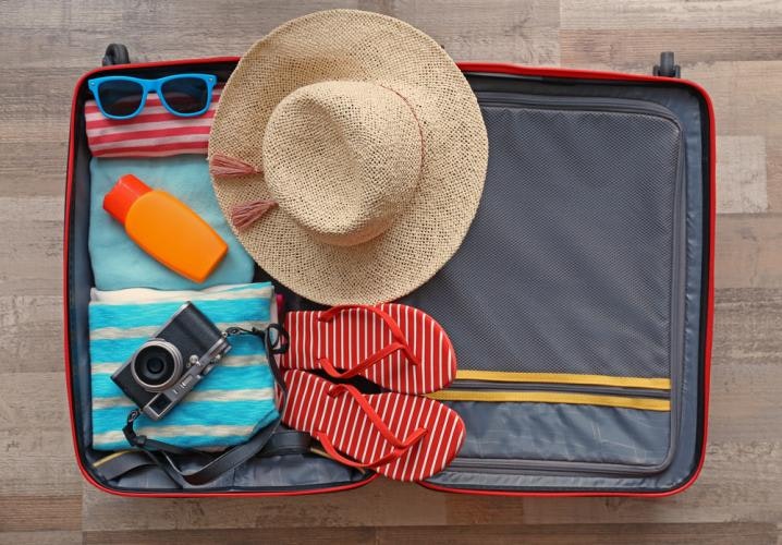 A view from above of an opened packed suitcase. The suitcase contains a straw hat with wide brim, red and white stiped flip flops, a camera, an orange bottle like a sunscreen bottle, a pair of blue-framed sunglasses, and a few towels.