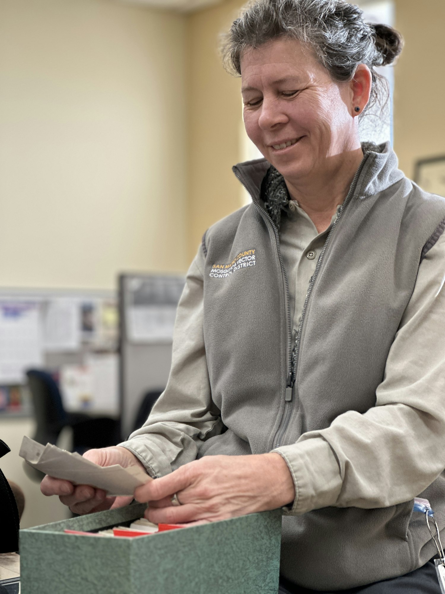 A person in a gray vest is smiling, holding papers from a green box.