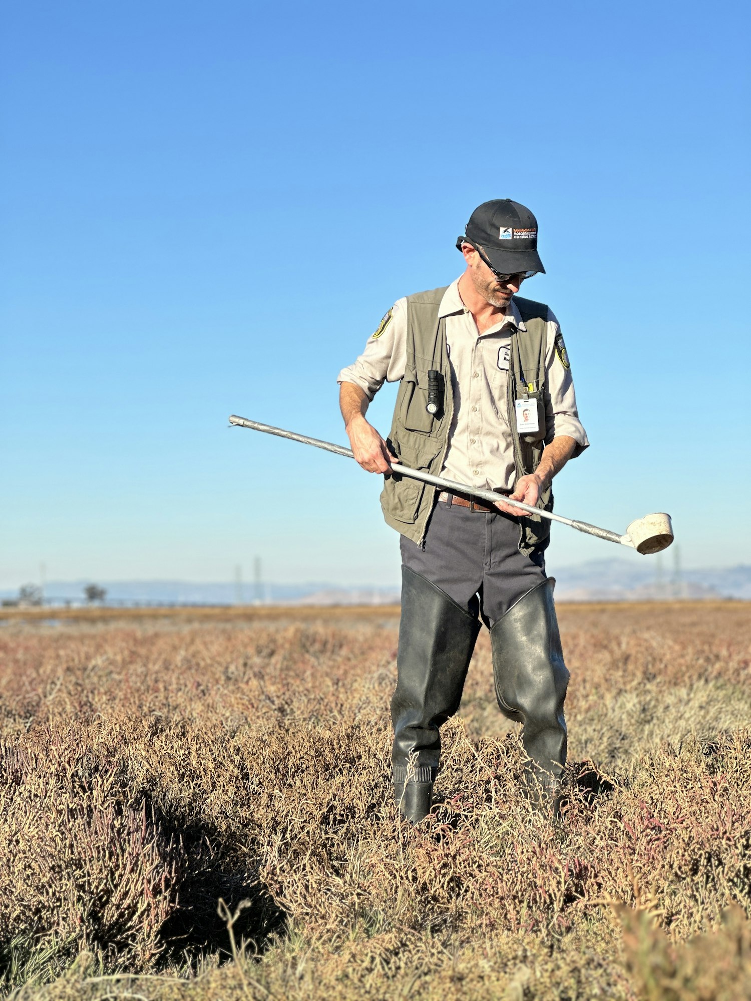 A person in a field wearing waders, holding a long pole, under a clear blue sky.