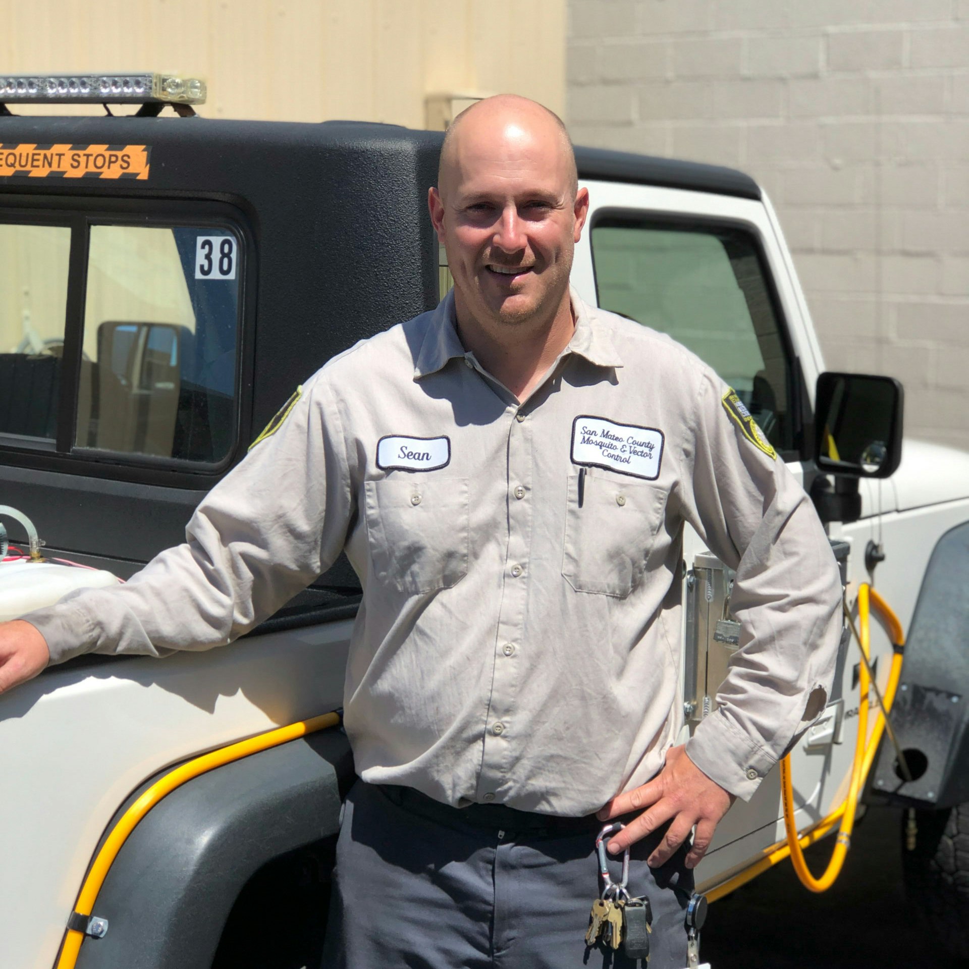 Smiling person wearing tan uniform shirt standing near a white Jeep with special lights and hoses attached.