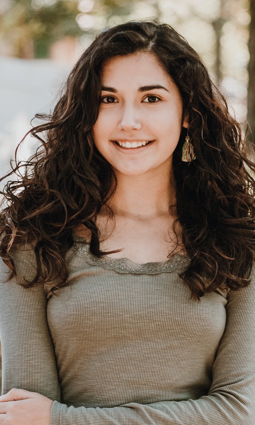 A person with curly hair smiling, wearing a green top, standing outdoors.