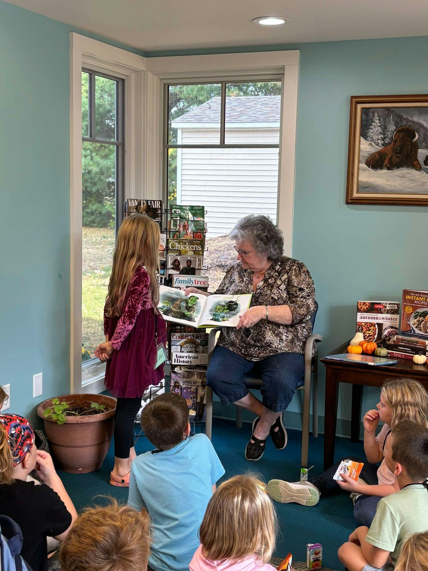 A woman is reading a book to children in a cozy room with a bison painting & magazines.