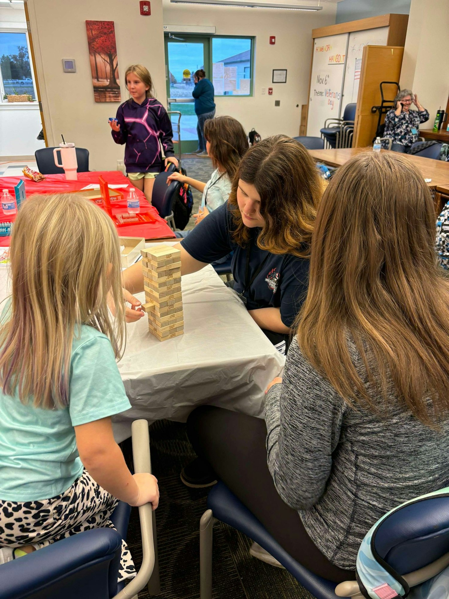 People playing Jenga at a table with onlookers in a room with autumn-themed decor.