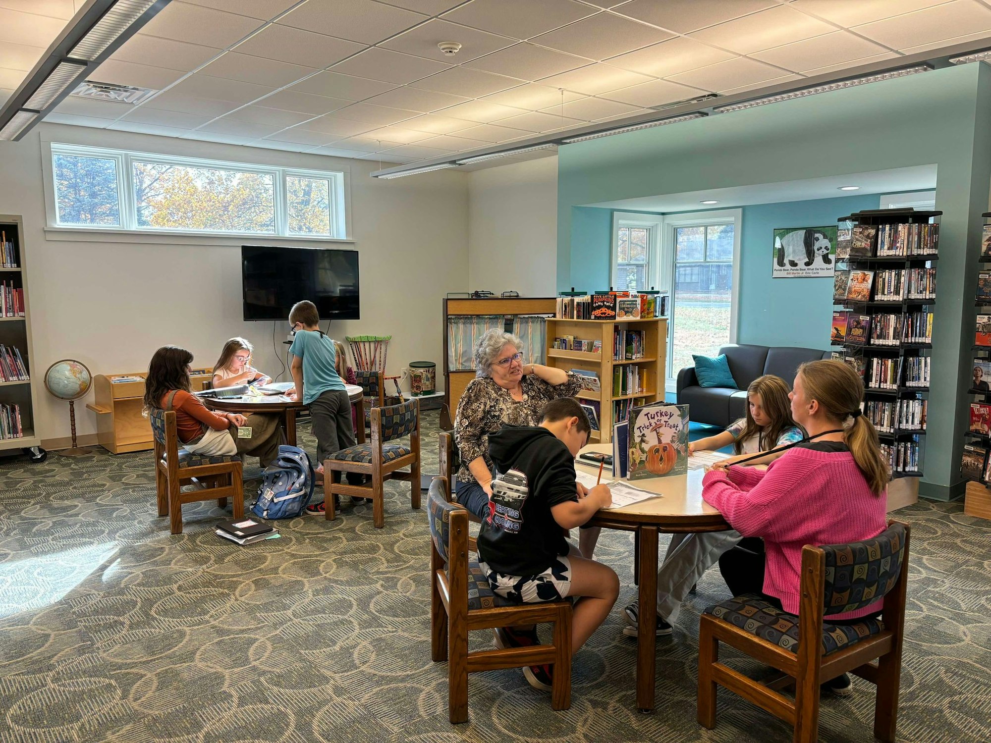 A group of children and adults in a bright library setting, some reading and some engaging in an activity at a table.
