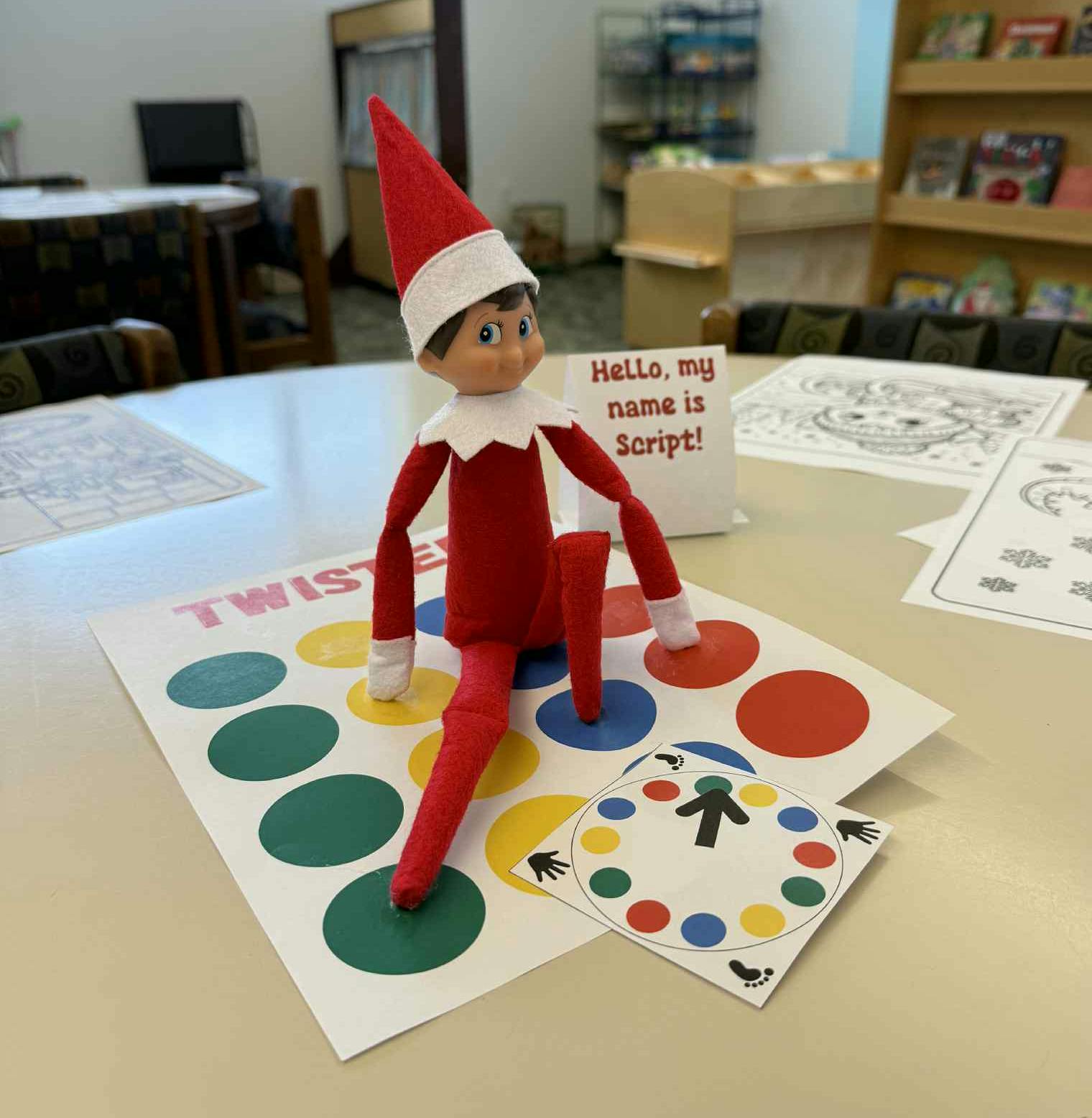 An elf doll named Script sits on a Twister game mat with a spinner, in a bright room decorated for fun.