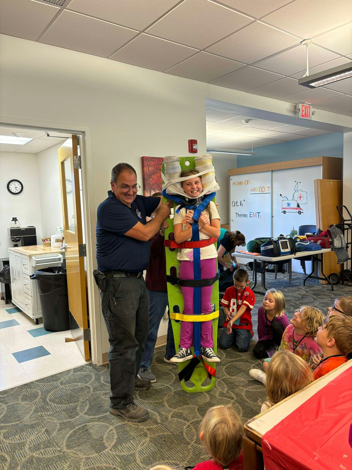 A group of kids watching as an adult wraps a child in foam pads and colorful straps, likely a fun learning activity.